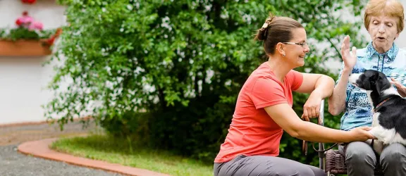 Mitarbeiterin der Rummelsberger Diakonie mit Hund und Seniorin im Rollstuhl