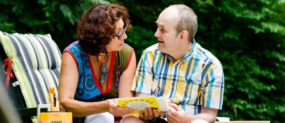Betreuerin sieht mit behinderten Mann auf einer Liege im Garten in ein Buch