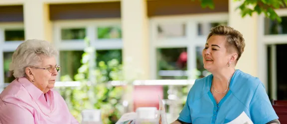 Seniorin mit Pflegefachkraft im Garten beim Zeitunglesen