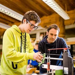 Ein junger Mensch steht an einer Maschine. Ein Lehrer schaut ihm zu.