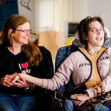 Eine Mitarbeiterin hält die Hand einer Frau im Rollstuhl. Beide lachen.