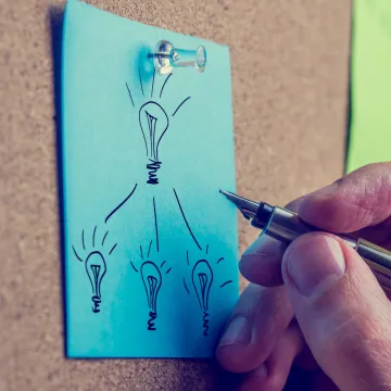 Eine Hand mit Stift malt Glühbirnen auf einen blauen Zettel