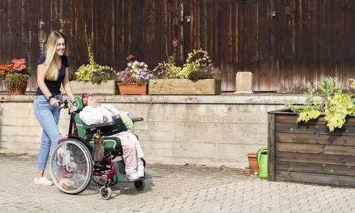 Mitarbeiterin schiebt eine Beschäftigte im Rollstuhl über den Hof