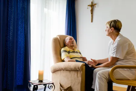 Ein alter Mann sitzt im Sessel. Gegenüber einer Pflegerin.